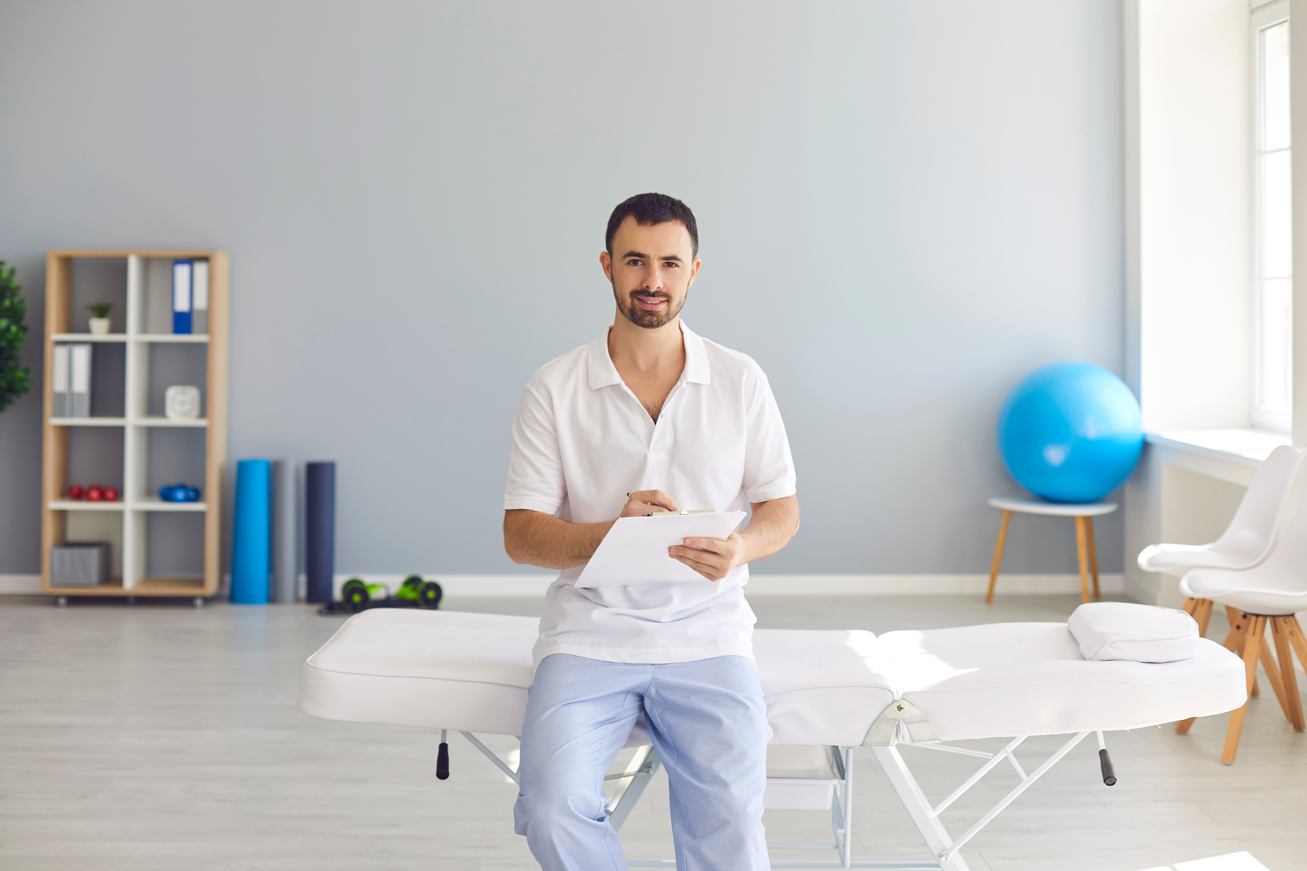 Young Male Doctor with Clipboard Sitting on Hospital Bed in Office of New Clinic